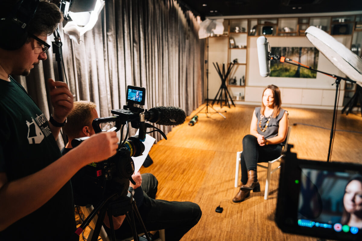 Production company crew behind a camera in a studio where a woman is being interviewed.
